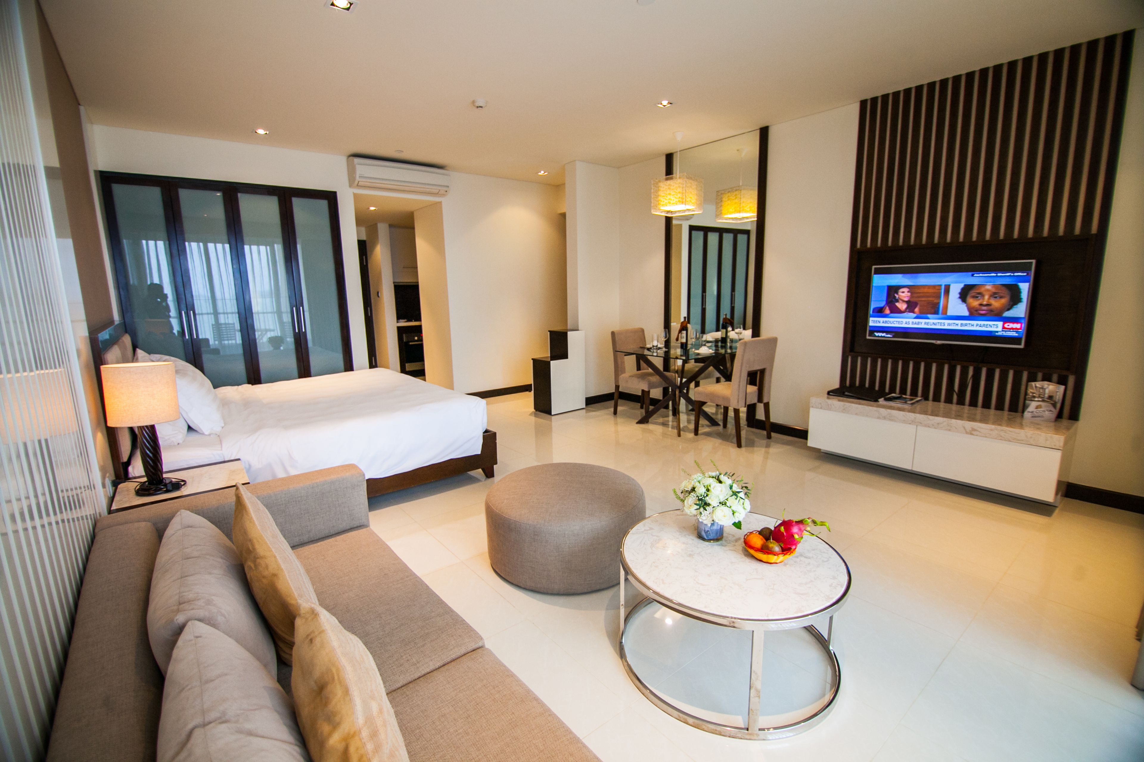 A modern hotel room with a bed, sofa, dining table, and a TV.