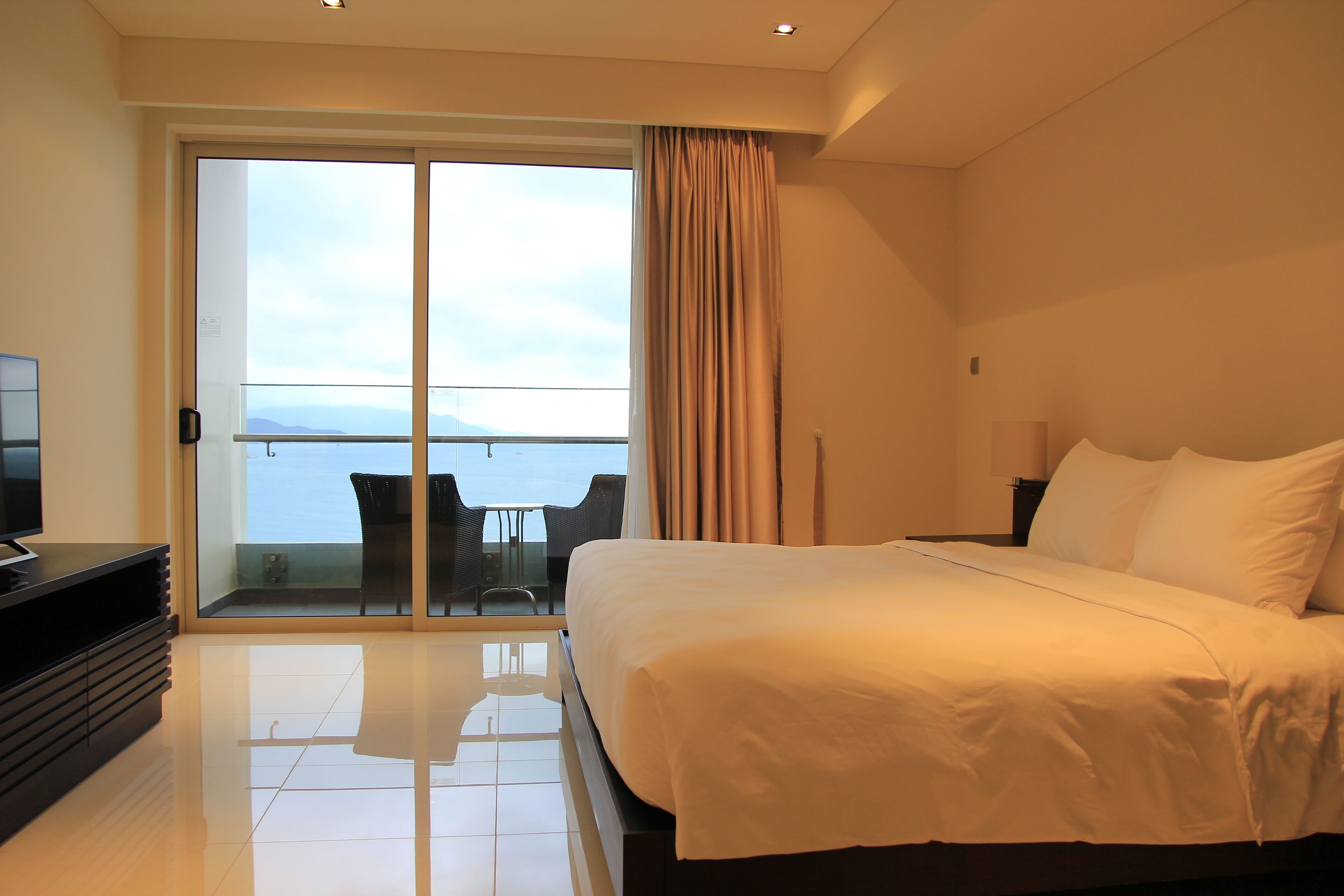 Un cuarto de hotel con una cama grande, televisión, balcón con mesa y sillas, y vista al mar.