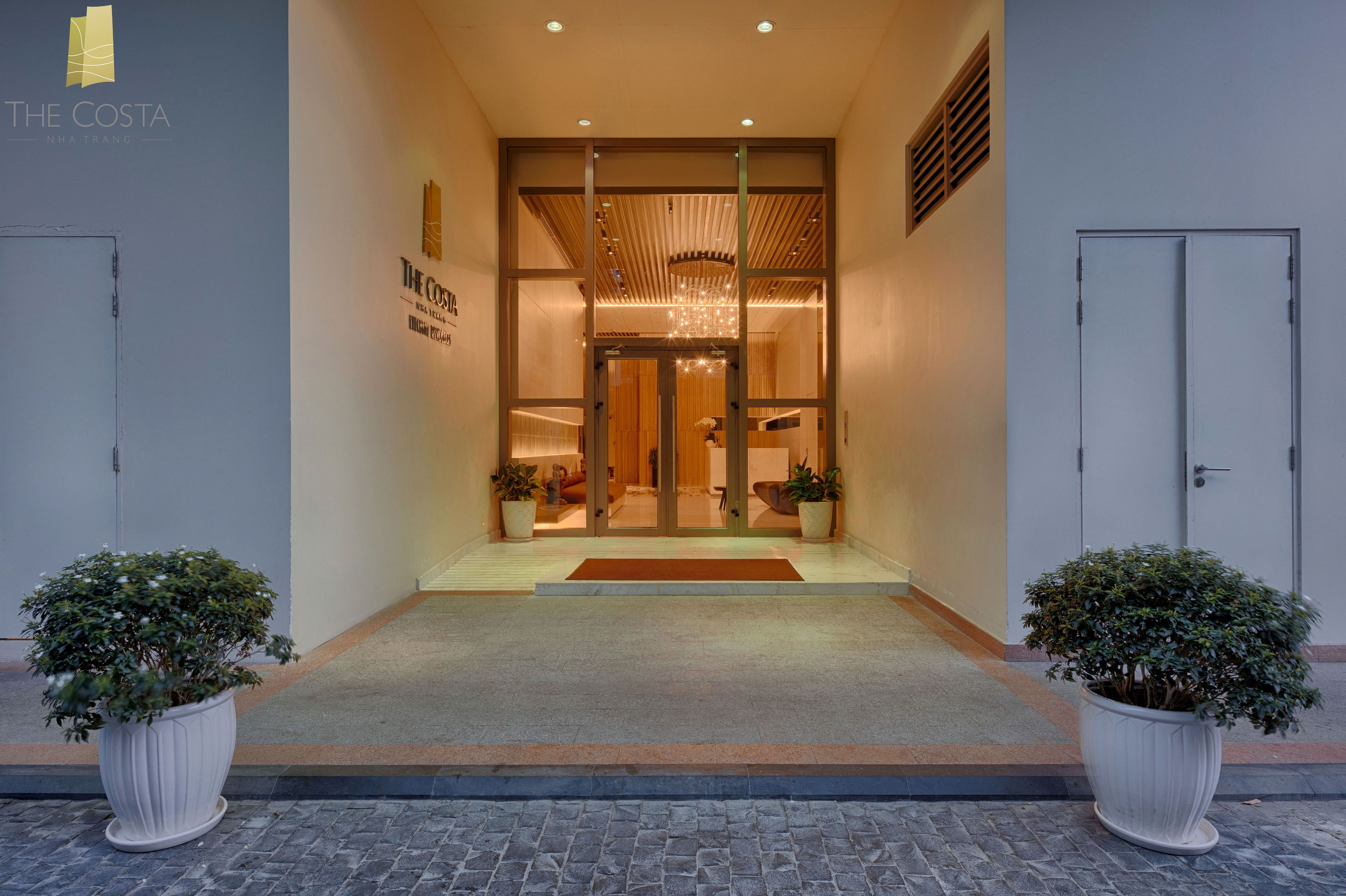 A hotel entrance with a modern design, featuring a glass door, a wooden ceiling, and potted plants on either side.