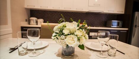 A dining table with a vase of white flowers, two wine glasses, and a plate in a modern kitchen with white cabinets and a microwave.