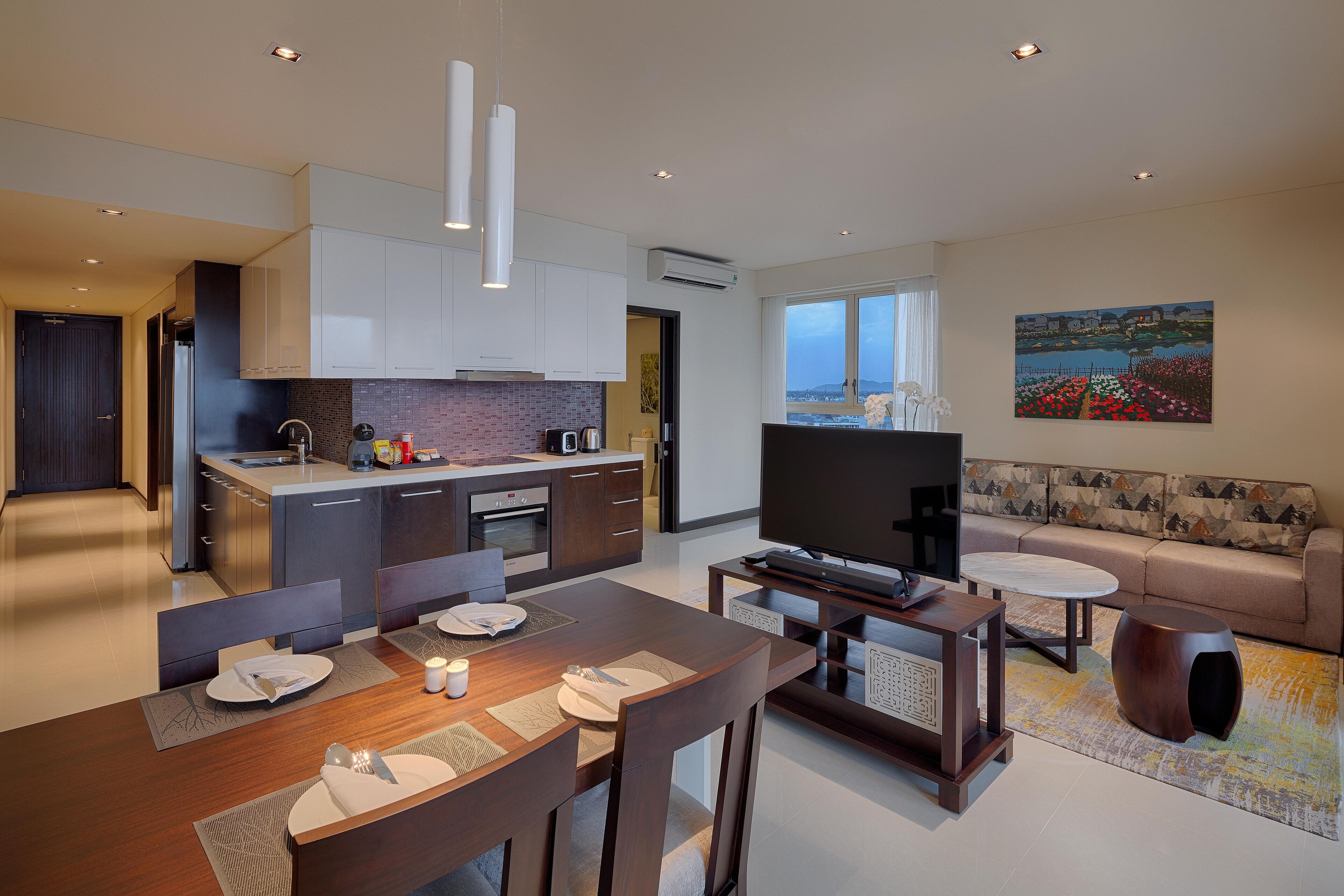 A modern kitchen with white cabinets, a dining area with a wooden table, and a living room with a sofa and a TV.