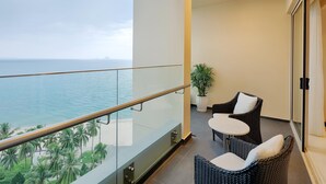 Un balcon doté d’une balustrade en verre, offrant une vue sur une plage et l’océan. On y trouve deux fauteuils en osier et une petite table sur laquelle est posée une plante en pot.