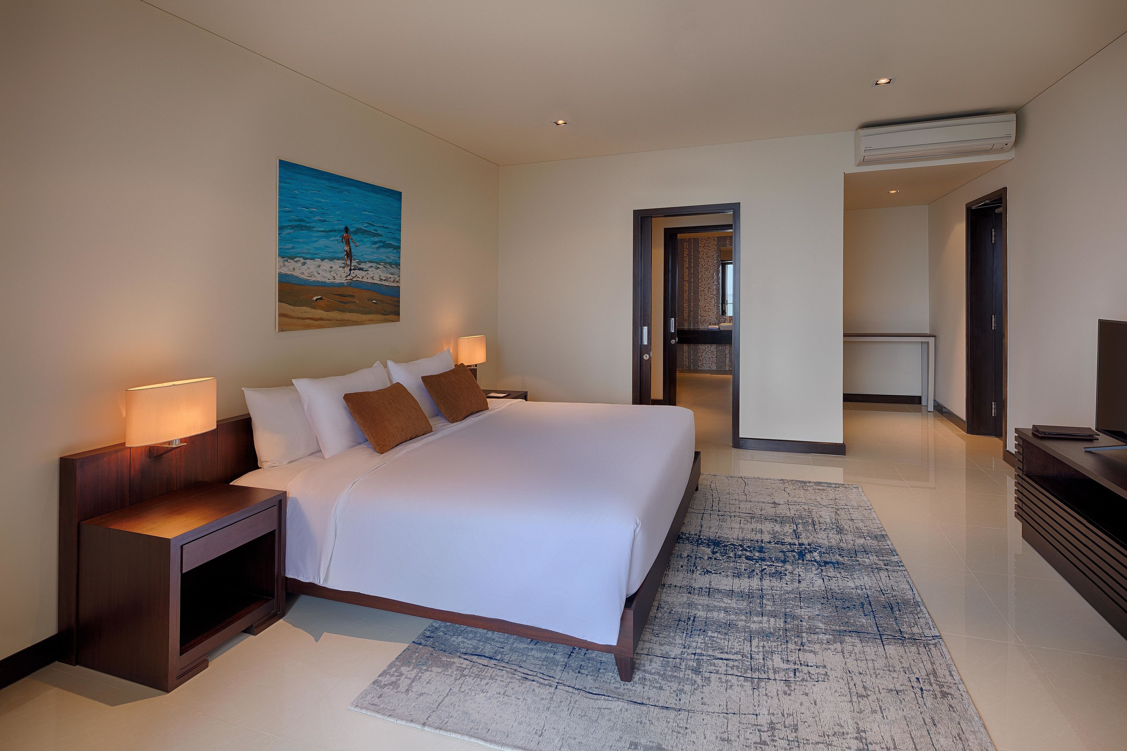 A modern hotel room with a large bed, bedside lamps, a painting of a beach scene, and a view of the hallway.