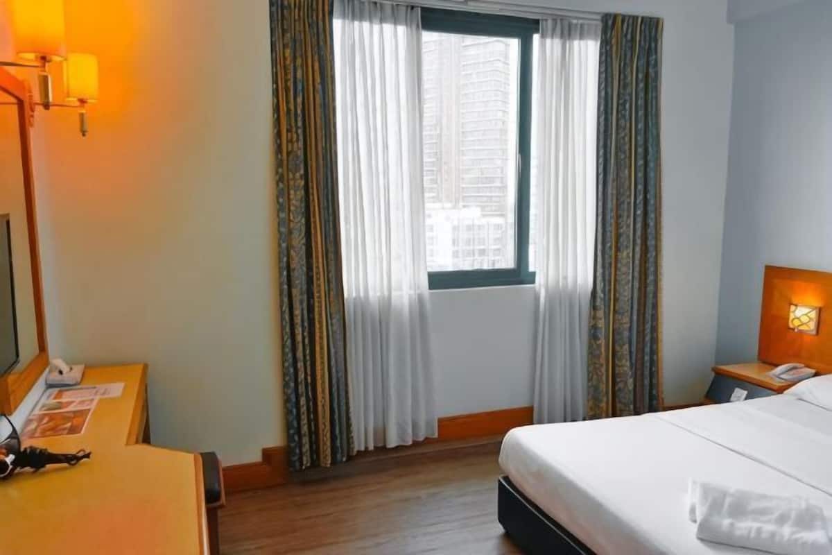 A hotel room with a bed, a desk with a lamp, a telephone, and a window with curtains.