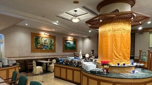 Un comedor de hotel con disposición tipo bufé, que incluye una isla central con comida y bebidas, y arreglos de asientos alrededor.