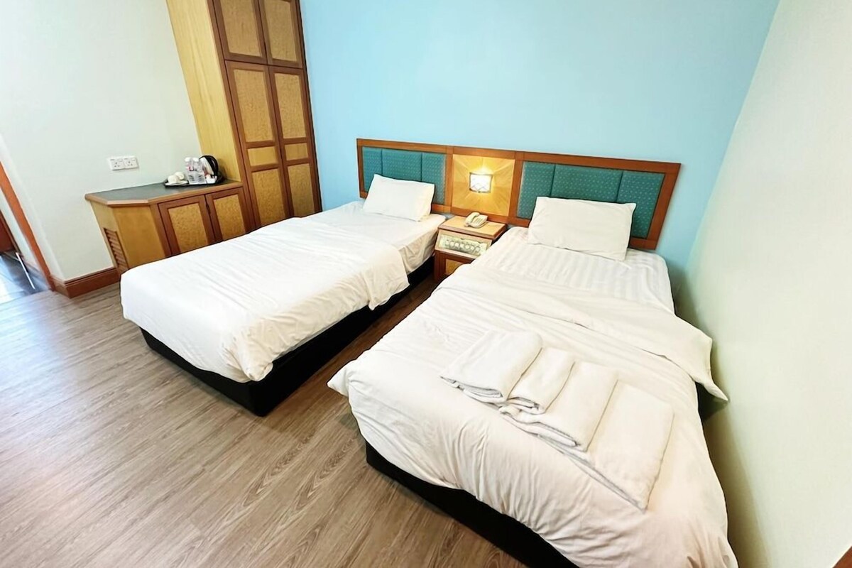 Habitación de hotel con dos camas, un armario de madera, una mesita de noche con lámpara y un pequeño escritorio con teléfono.