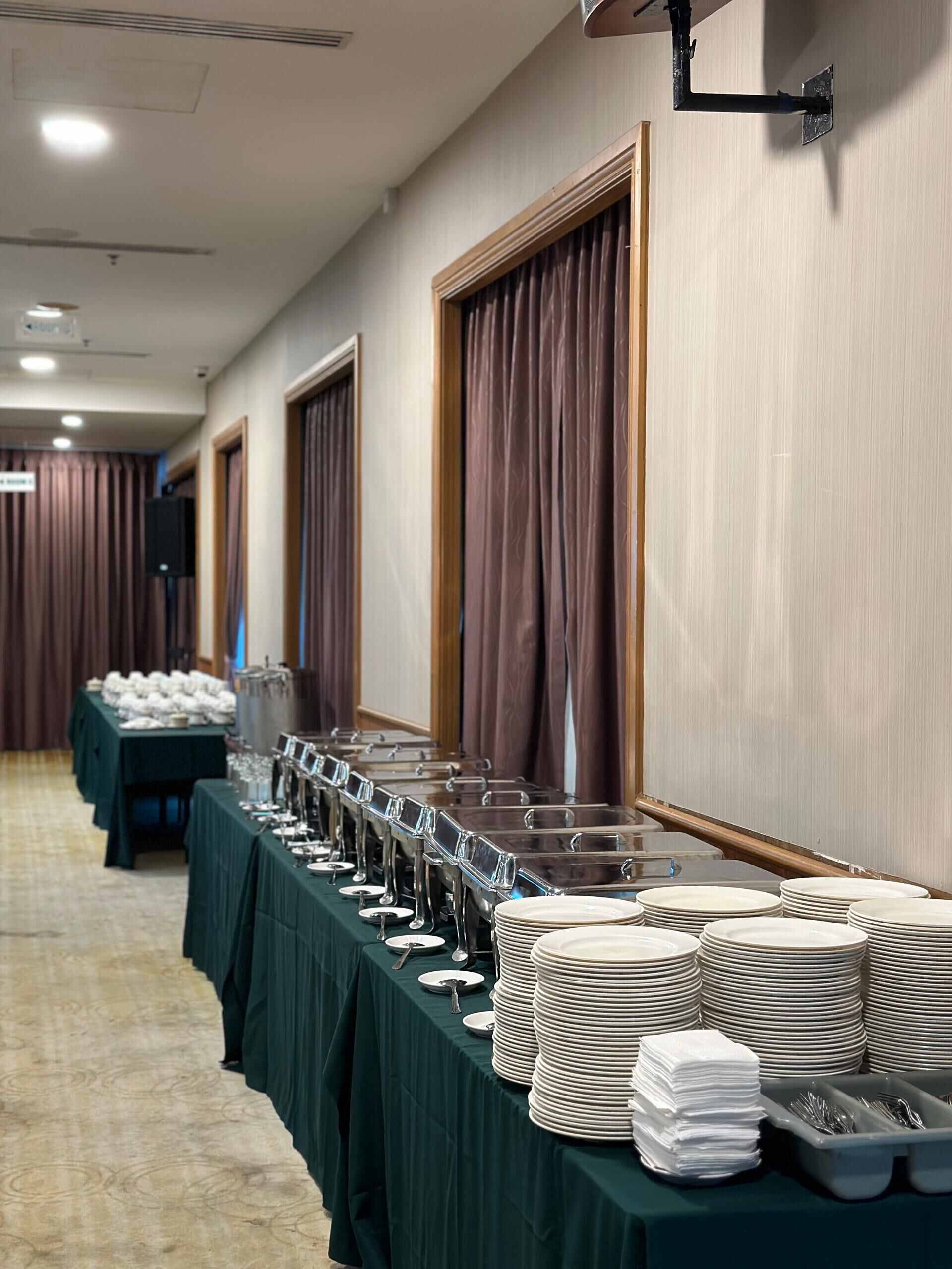 A banquet hall with long tables covered in green tablecloths, equipped with glassware and stacked plates.