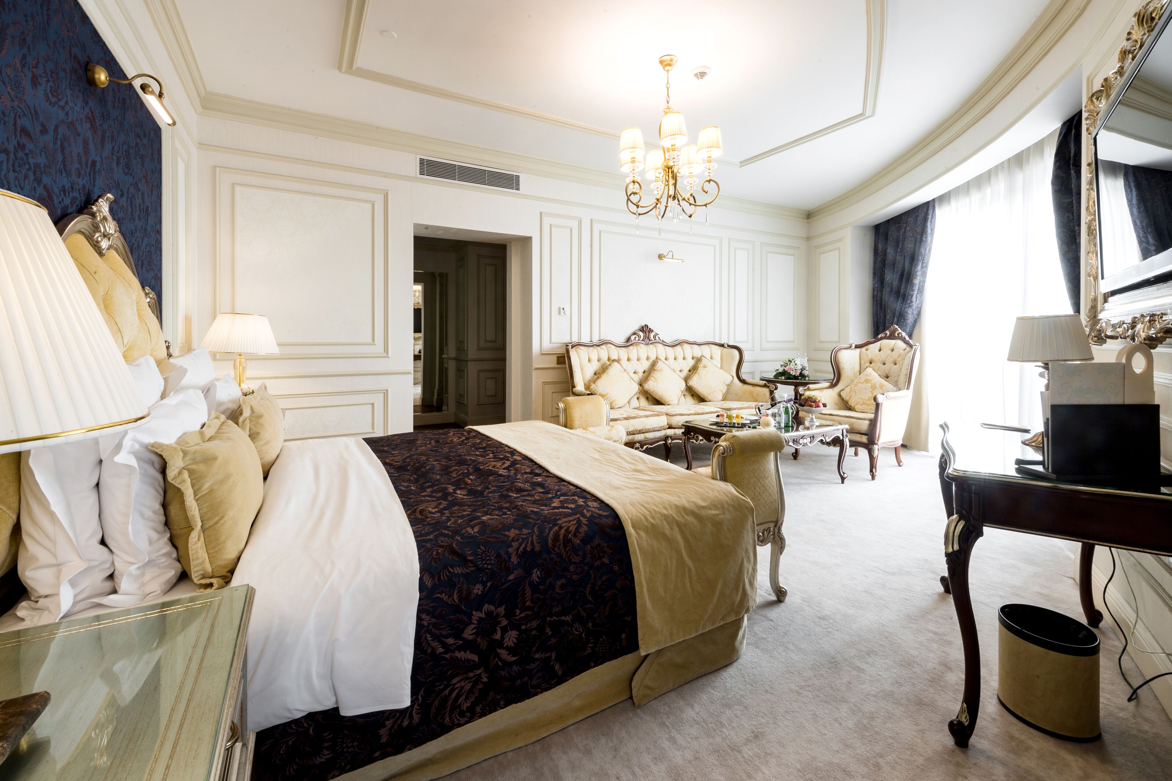 Amplia habitación de hotel con una cama grande, una zona de estar con sofá y sillones, una mesa de comedor y una televisión.