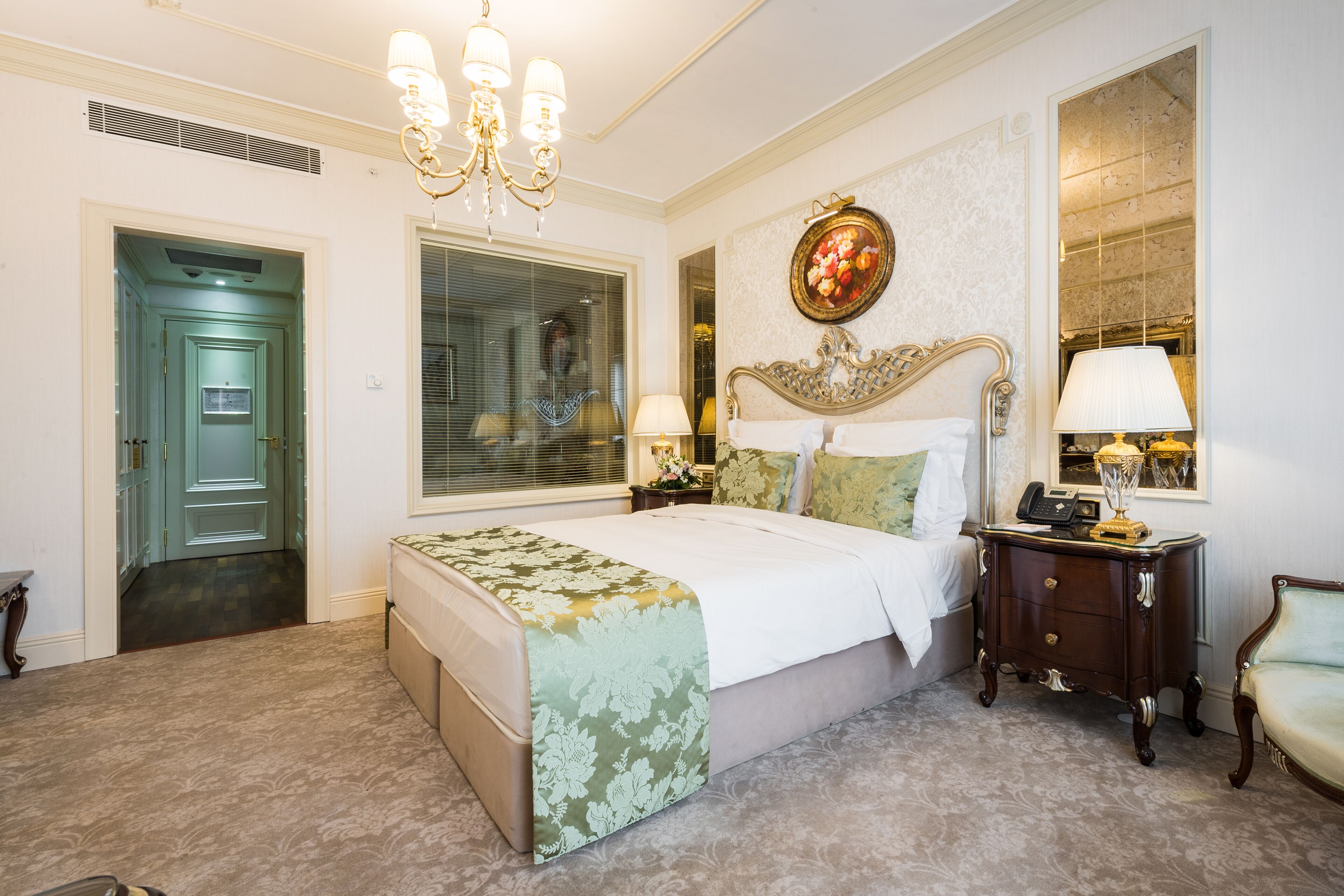 A bedroom with a large bed, a nightstand with a lamp, a chair, a mirror, and a door leading to another room.