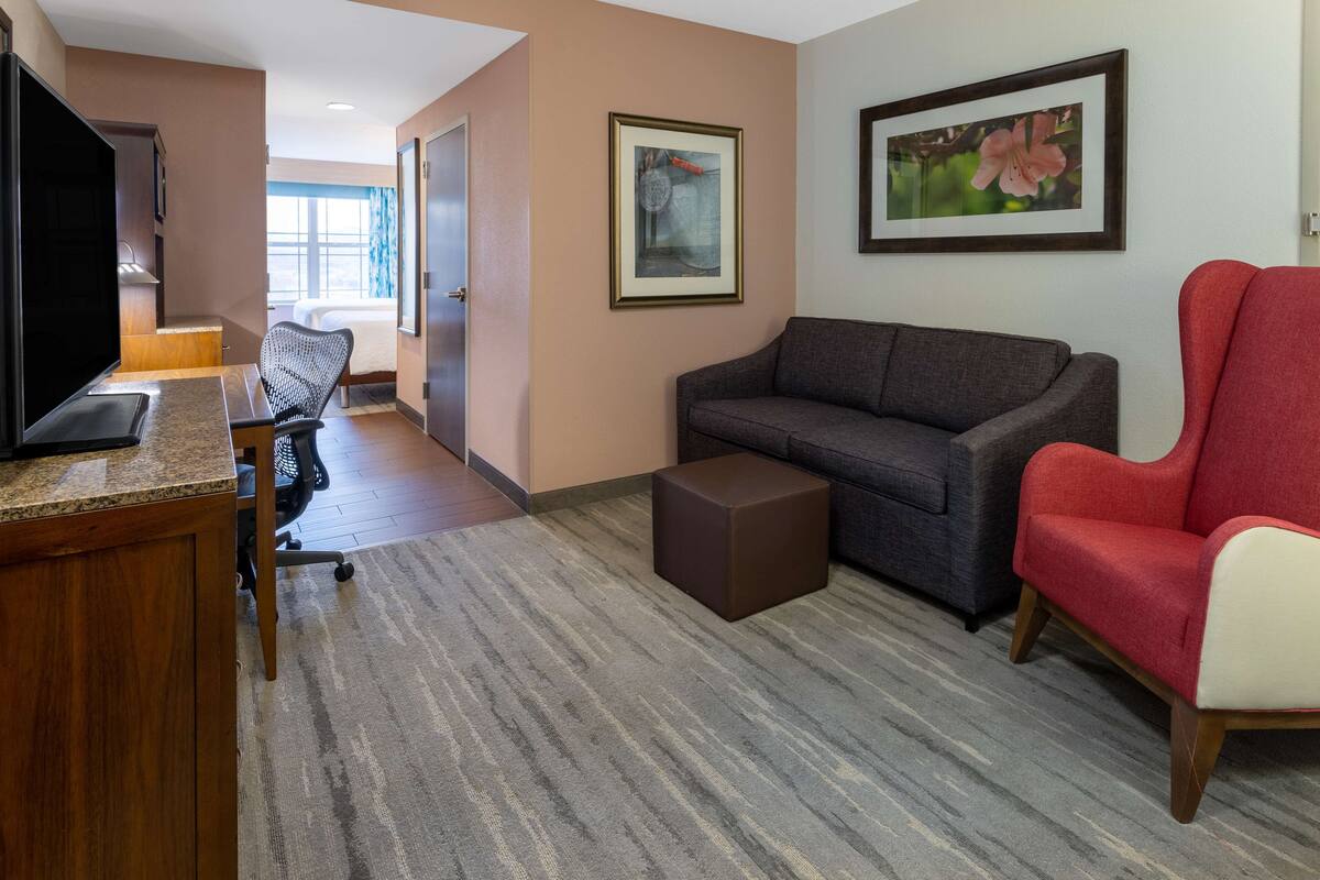 A hotel room with a television, a gray sofa, a red armchair, a small ottoman, and a framed picture on the wall.