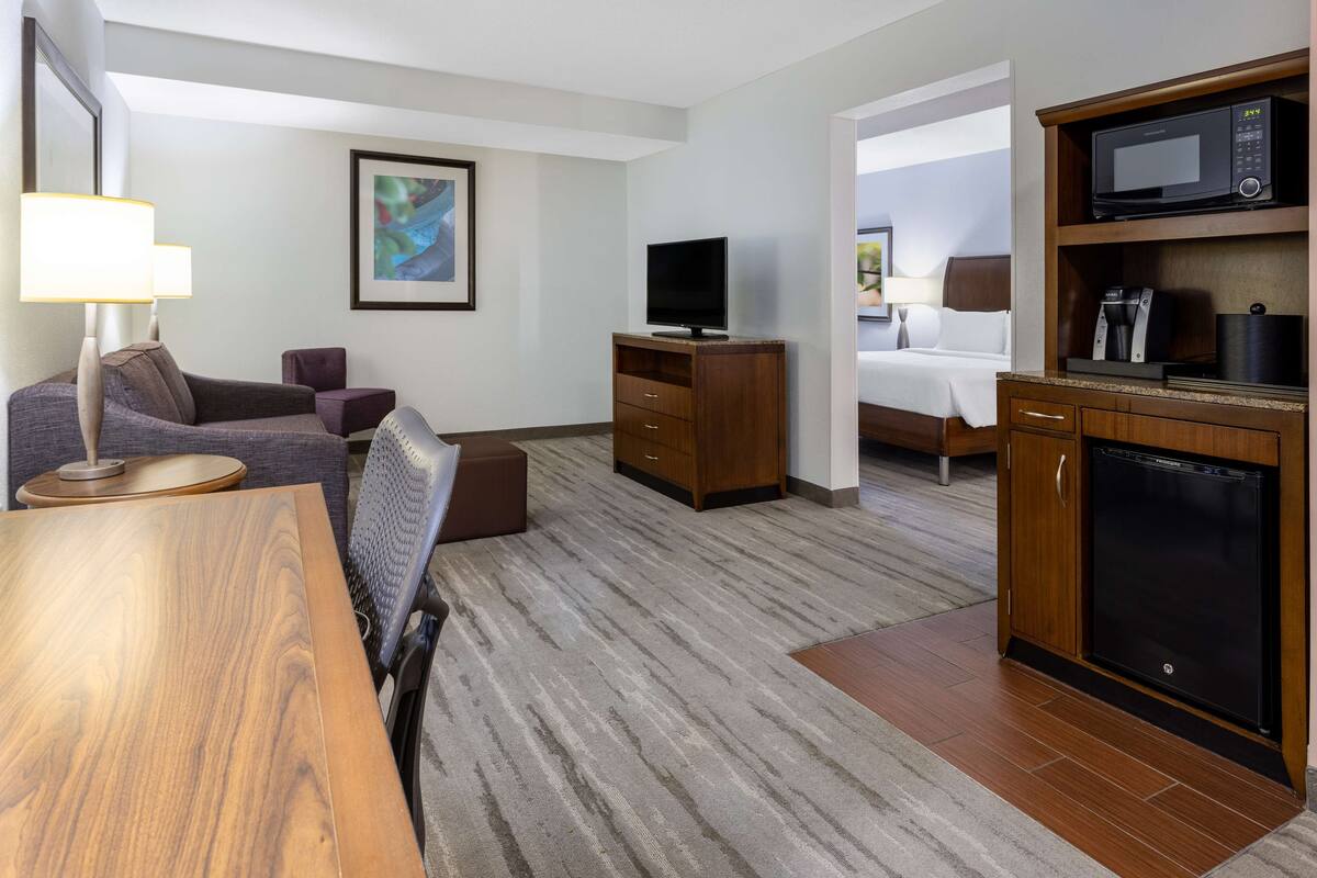 A hotel room with a wooden dining table, a sofa, a television on a wooden stand, a microwave, and a refrigerator.