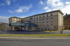 A multi-story hotel with a central entrance, a parking lot, and a hilly landscape in the background.