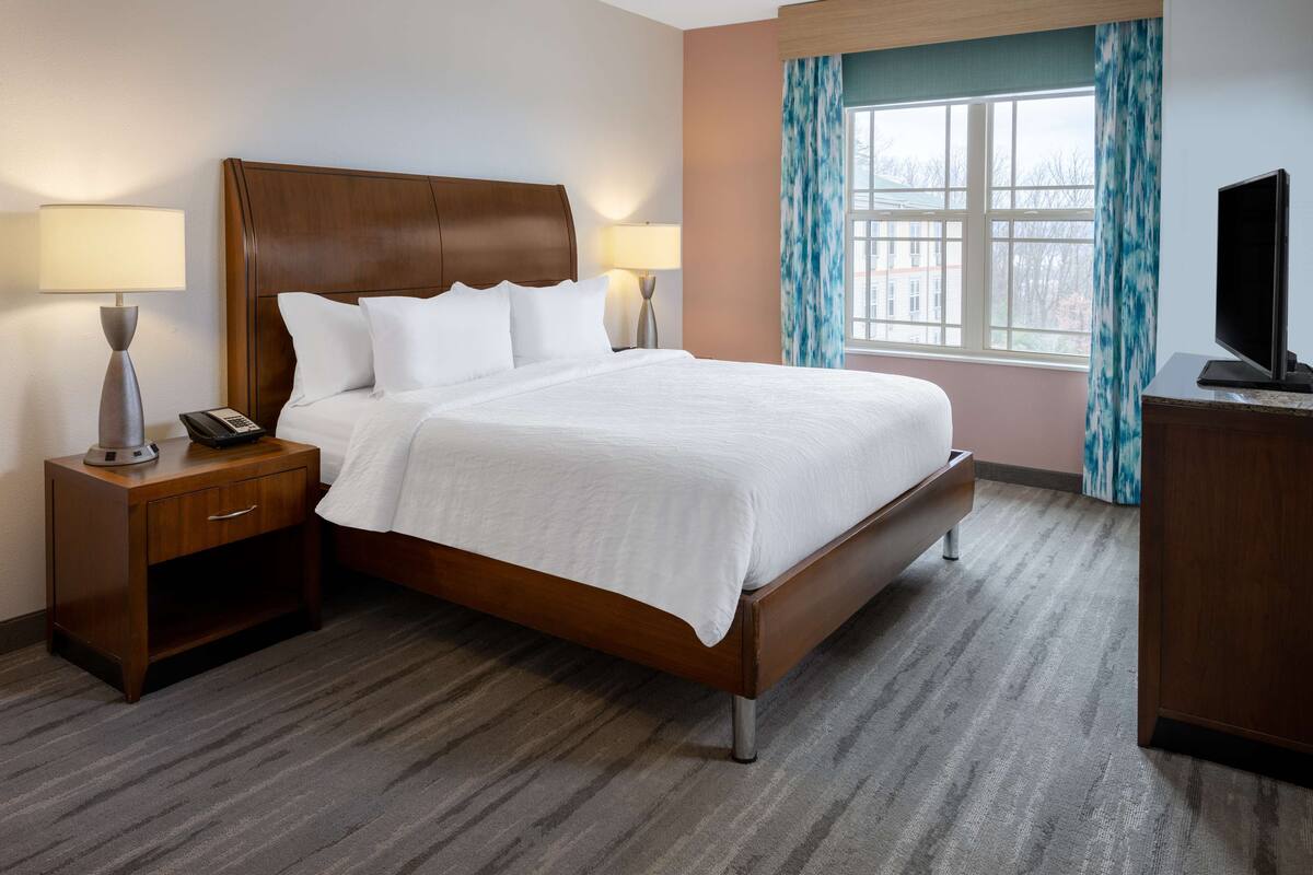 A neatly made bed with a wooden headboard, two bedside tables with lamps, a television, and a window with curtains.
