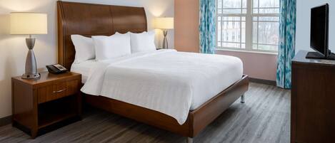 A neatly made bed with a wooden headboard, two bedside tables with lamps, a television, and a window with curtains.