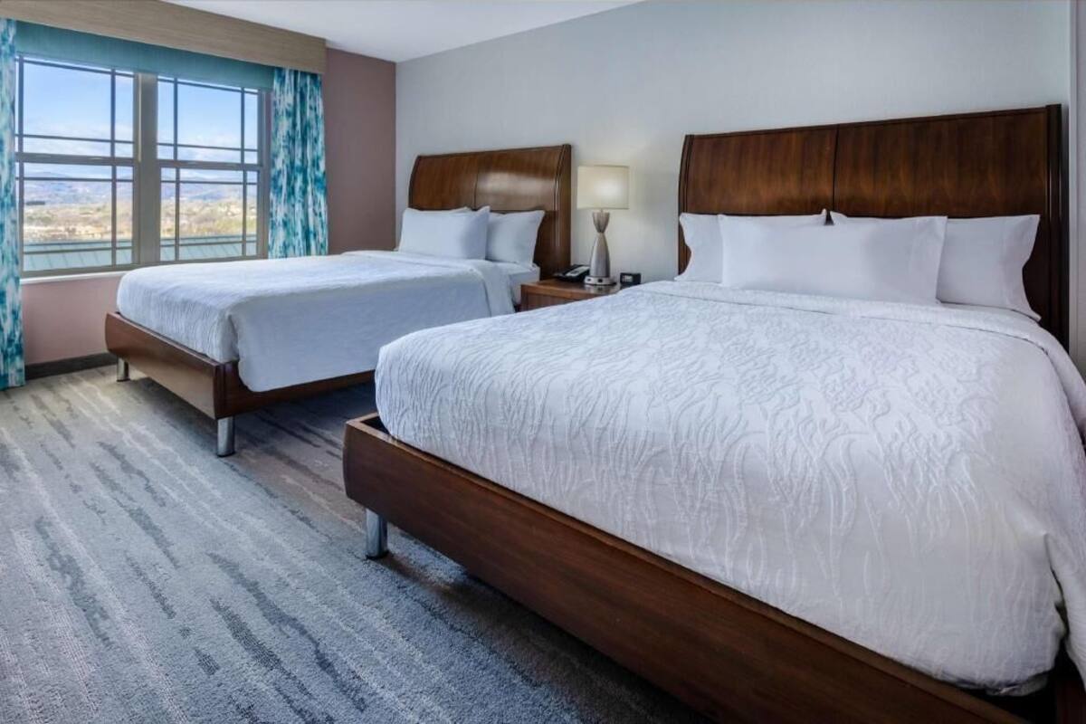 A hotel room with two beds, a large window with curtains, a lamp, and a view of the landscape.
