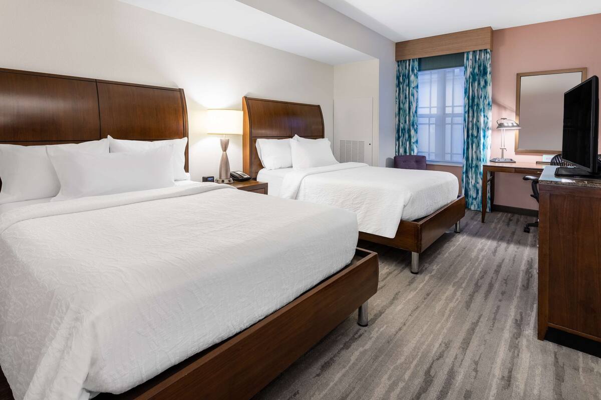 A hotel room with two beds, a desk, a chair, a TV, and a window with curtains.