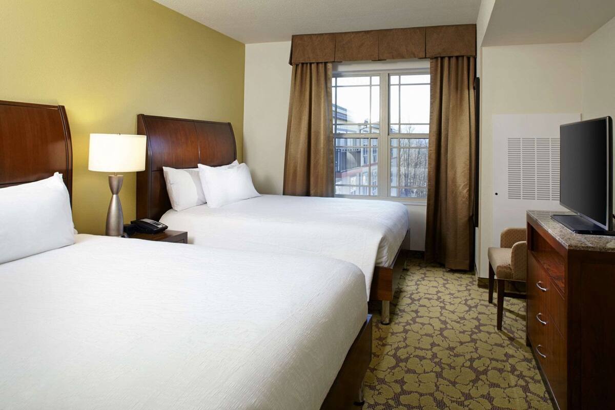 A hotel room with two beds, a desk with a lamp, a television, and a patterned carpet.