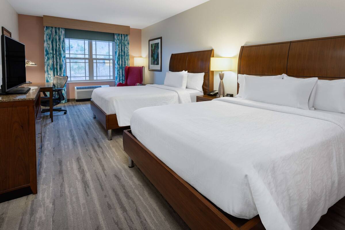 A hotel room with two beds, a desk with a computer, a chair, a TV, and a window with curtains.