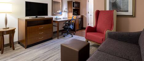 Habitación de hotel con televisor de pantalla plana, escritorio de madera, silla de oficina, sillón rojo, sofá gris y un pequeño reposapiés.