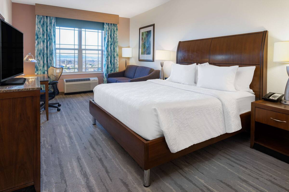 A hotel room with a large bed, a desk with a TV, a chair, and a window with curtains.