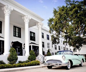 Un edificio clásico blanco con columnas y contraventanas negras, un coche antiguo estacionado al frente, y follaje cuidado.