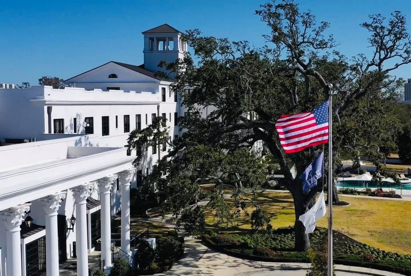 A white building with a tower, surrounded by trees and a flagpole with flags.