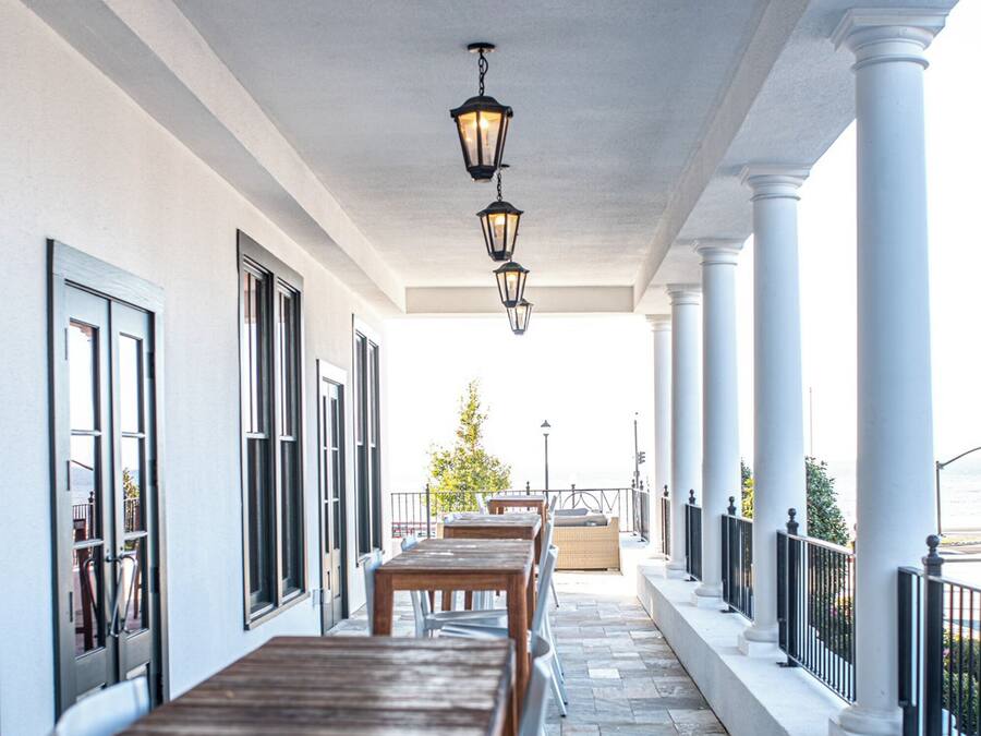 A covered outdoor patio with a row of tables and chairs, white columns, and hanging lanterns.