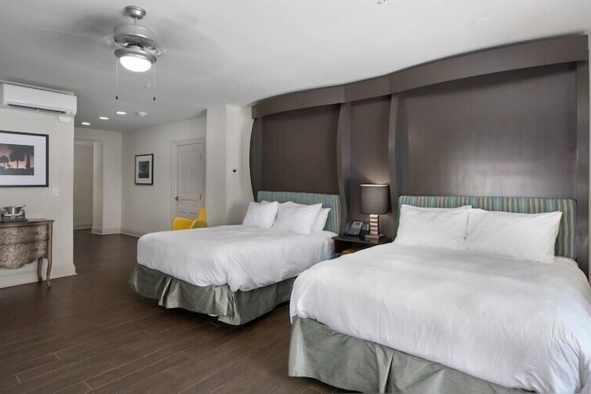 Habitación de hotel con dos camas, ventilador de techo, aire acondicionado, mesita de noche con lámpara y un cuadro en la pared.
