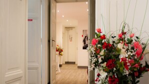 A hallway with wooden flooring, white walls, and a door leading to another room. There is a potted plant with red and white flowers on a table. - Relais Cola di Rienzo (Rome)