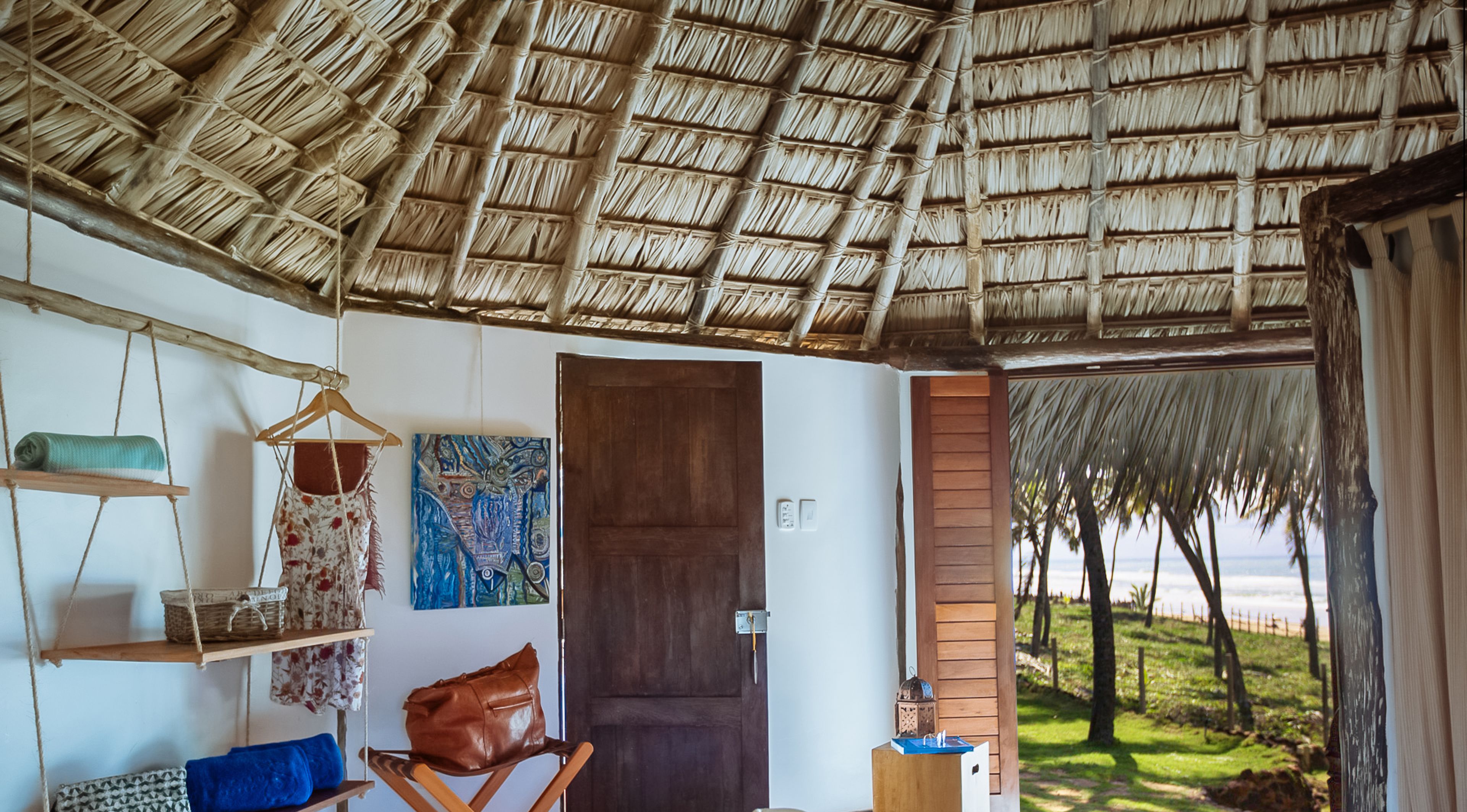 A room with a thatched roof, a wooden door, a hanging chair, a shelf with a basket, a painting, and a view of the beach.