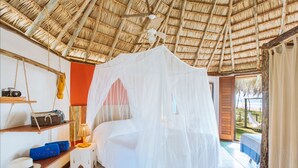 Une chambre avec un lit recouvert d’une moustiquaire, une étagère en bois sur laquelle se trouvent un appareil photo et un panier, et une vue sur la plage par une porte ouverte.