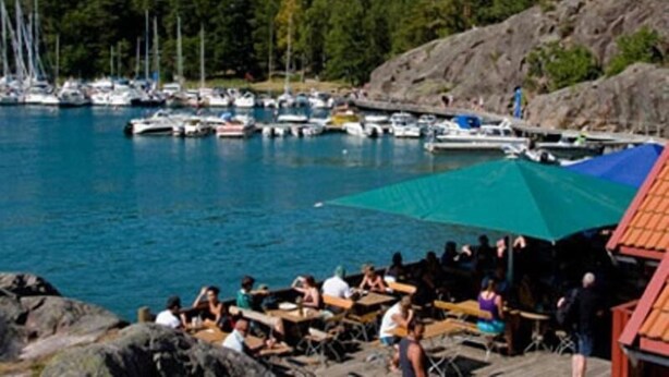 Un puerto deportivo con barcos amarrados, una costa rocosa y personas sentadas en mesas bajo una tolda.