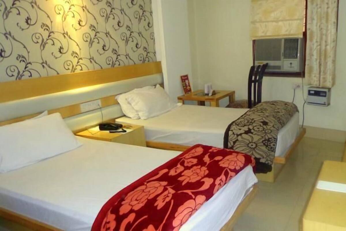 Ein Hotelzimmer mit zwei Betten, einem kleinen Tisch, einem Nachttisch, einem Fenster mit Vorhängen und einer Klimaanlage.