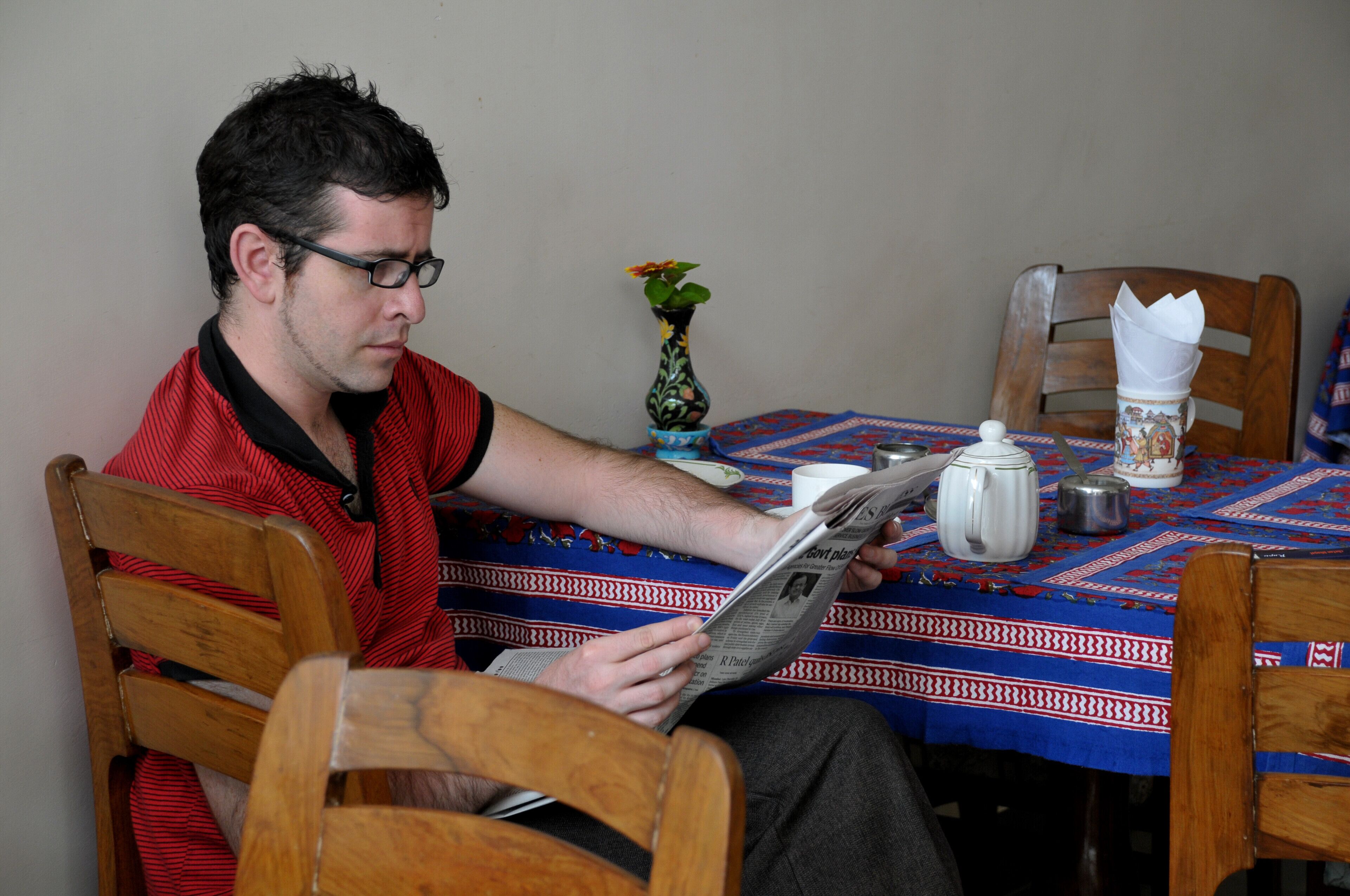 Una persona leyendo un periódico en una mesa de madera con mantel estampado, un jarrón con flores y un hervidor.