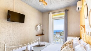 A hotel room with a bed, a mounted TV, a balcony with a view, and a small table with a lamp. - Hotel Ariadne (Skopelos)
