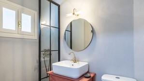 A bathroom with a white sink, a round mirror, a toilet, and a glass shower enclosure. - Hotel Ariadne (Skopelos)
