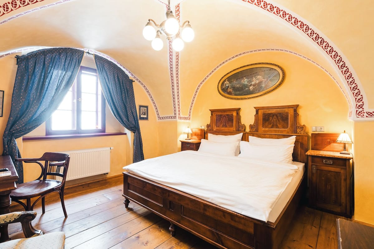 A bedroom with a wooden bed, a chair, a desk, a window with curtains, a radiator, and a chandelier.