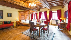 Quarto com cama de dossel, vigas de madeira no teto, mesa de jantar com cadeiras e cortinas vermelhas.