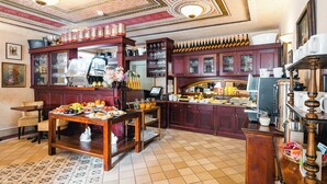 Cozinha de hotel tradicional, com armários de madeira, piso ladrilhado e diversos alimentos e bebidas expostos em mesas.