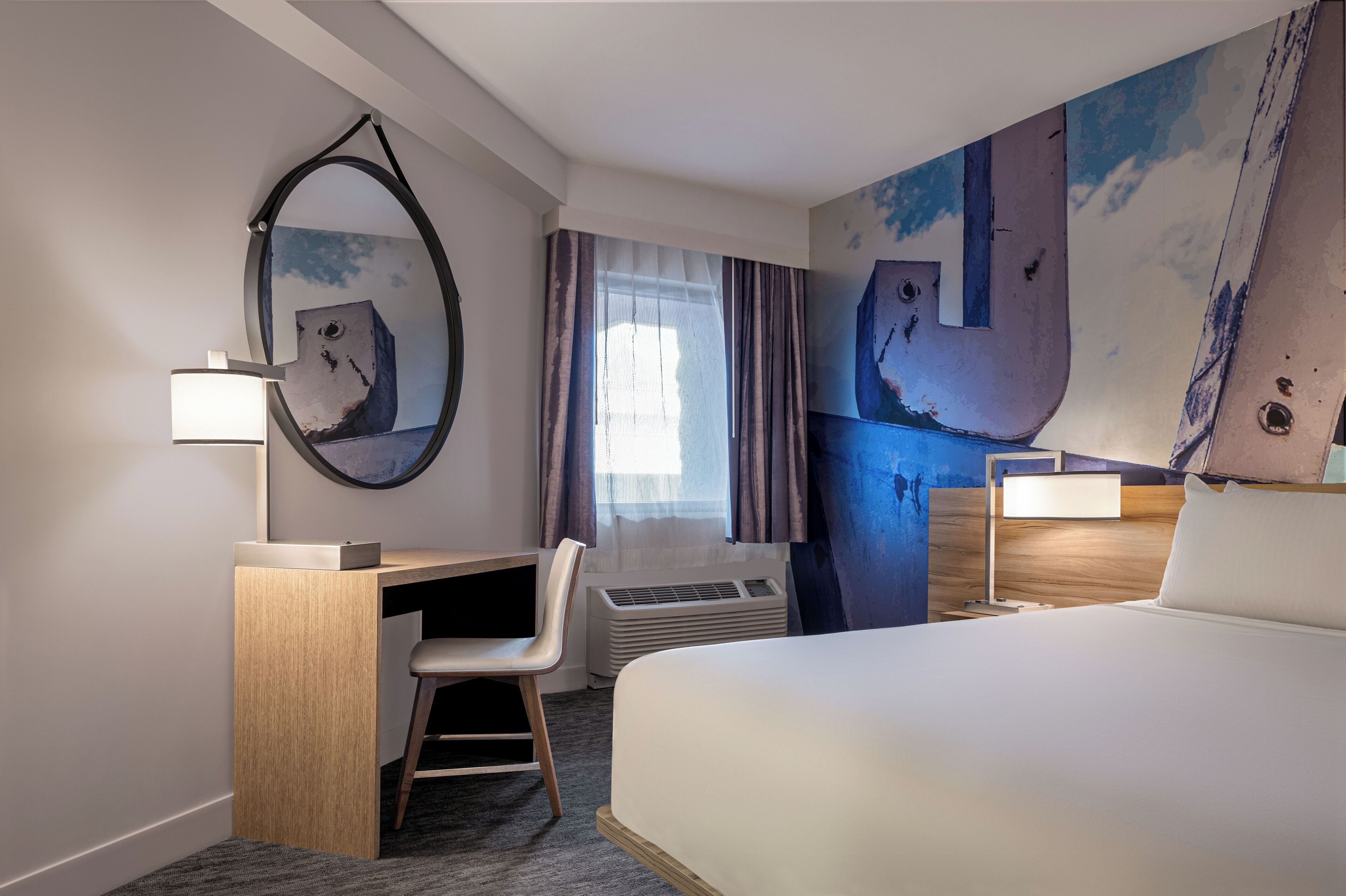 Een hotelkamer met een bed, een bureau met stoel, een bureaulamp, een grote spiegel, een raam met gordijnen, en een muur met een groot abstract schilderij.