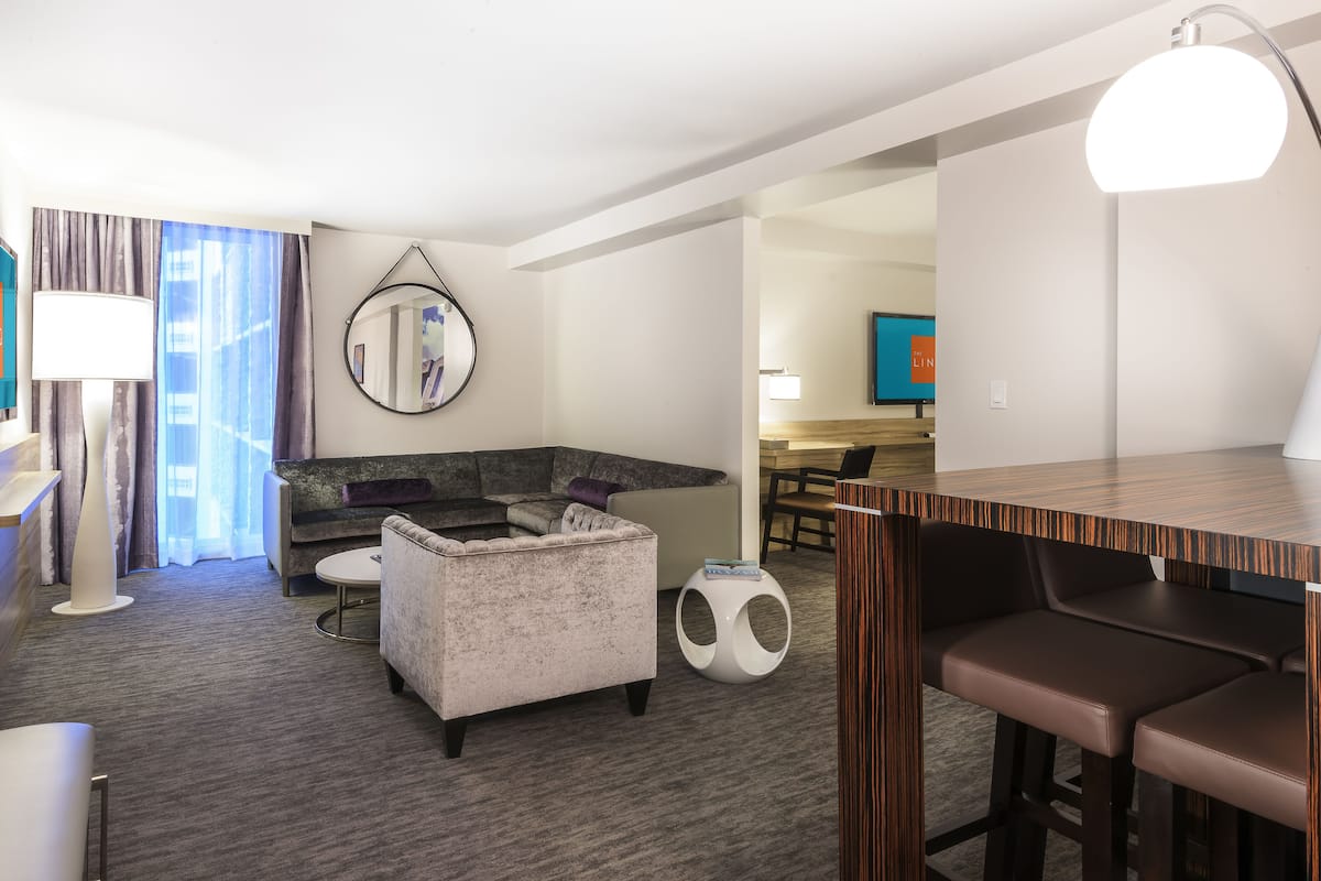 A modern hotel room with a grey sofa, a round coffee table, a small ottoman, a wooden bar area, and a TV mounted on the wall.