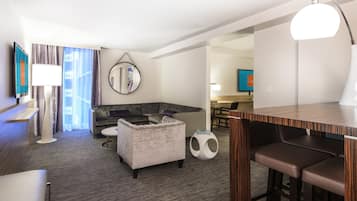 Una habitación de hotel moderna con un sofá gris, una mesa redonda, un pequeño otoman, una zona de bar de madera y un televisor montado en la pared.