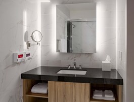 Un baño moderno con pared de azulejos de mármol, lavamanos con encimera negra, armarios de madera, espejo y ducha de mano.