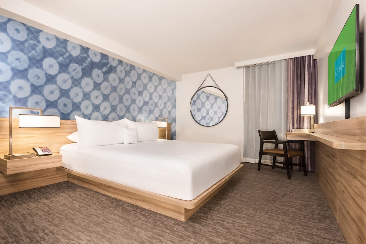 A hotel room with a large bed, a desk, a chair, a TV, a lamp, and a patterned wall.