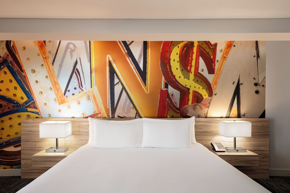A hotel room with a large bed, two bedside tables with lamps, and a colorful abstract mural on the wall.