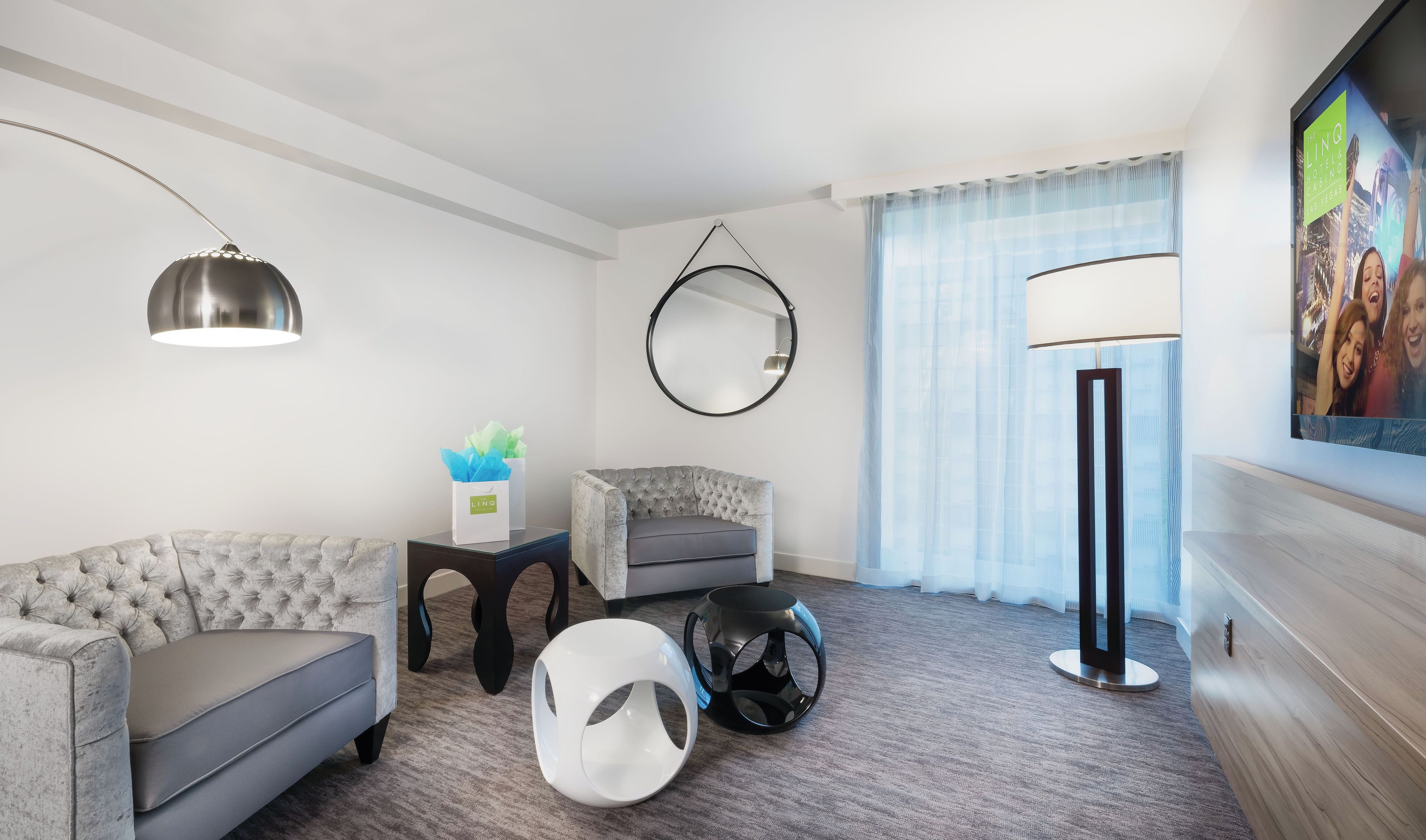 Una moderna camera d'albergo con un divano grigio trapuntato, uno specchio rotondo, una lampada da terra e un televisore a schermo piatto.
