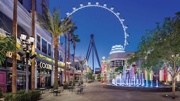 A nighttime view of a street with a large Ferris wheel in the background, palm trees lining the sidewalk, and a variety of storefronts including a visible cocktail bar.