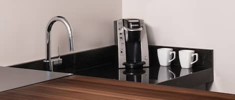 Una cocina moderna con encimera negra, un lavamanos de acero inoxidable, una cafetera y dos tazas blancas.