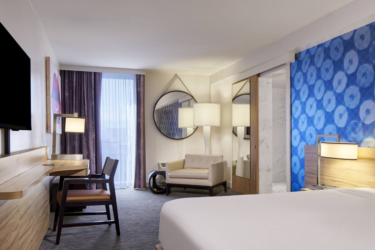 A hotel room with a bed, a desk, a chair, a TV, and a large window with curtains.