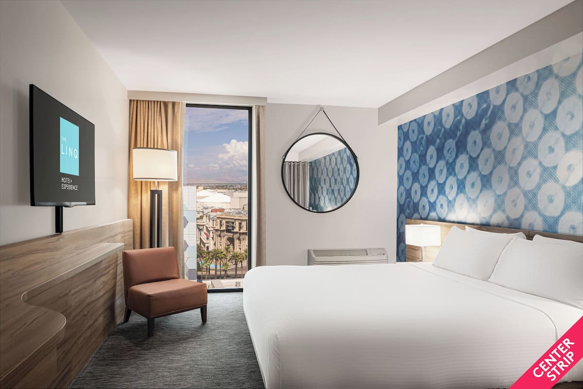 A hotel room with a large bed, a seating area, a TV, and a view of the city.